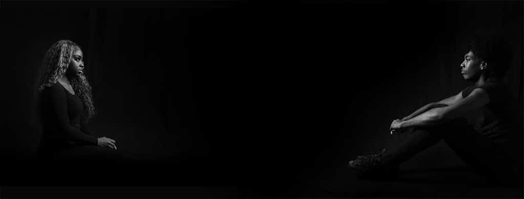 Two women sit facing each other in a dark room, both illuminated by soft light. One sits cross-legged with her arms on her knees, the other sits with hands on her lap. They are surrounded by deep shadows.