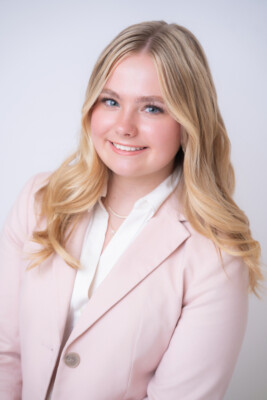 A young woman with long blonde hair, wearing a light pink blazer over a white blouse, smiles at the camera against a plain light background.