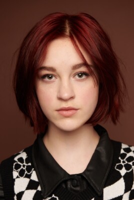 A young person with short, straight red hair and fair skin looks directly at the camera. They wear a black-and-white patterned sweater over a black collared shirt, with a neutral brown background.